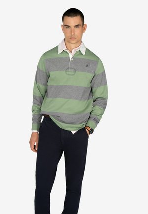 Camisa polo de mangas largas en rayas verdes y grises, con un color blanco, logotipo bordado y un corte relajado. Combinada con pantalones oscuros.