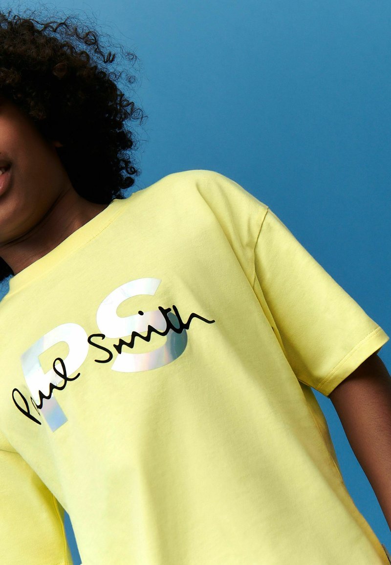 Camiseta de algodón amarilla con un logo en cursiva negro de "Paul Smith" y una impresión translúcida de "PS" en el frente. Manga corta, corte relajado.