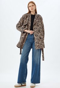 Abrigo de estampado de leopardo con cintura ceñida, corte oversized, mangas anchas y detalles de botones, combinado con jeans de pierna ancha en azul y zapatillas blancas.