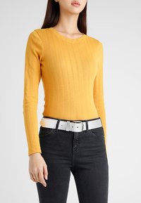 Haut jaune à manches longues et à côtes avec un col rond, associé à un jean noir et une ceinture blanche texturée avec une boucle en métal argenté.