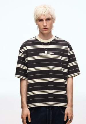 Junge Person mit kurz gebleichtem Haar, die ein schwarz-beiges gestreiftes T-Shirt mit dem Schriftzug "OVRNRM" und eine Halskette mit Anhänger trägt.