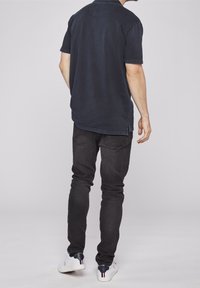 Męska granatowa koszulka polo z krótkim rękawem, klasycznym kołnierzem i rozcięciami po bokach, zestawiona z czarnymi dopasowanymi jeansami i białymi trampkami.