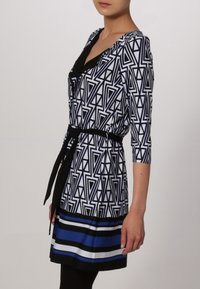 Czarna i biała sukienka we wzory geometryczne z rękawami o długości trzy czwarte, z paskiem w talii oraz niebiesko-czarnym paskiem na dole. Gładka faktura materiału.