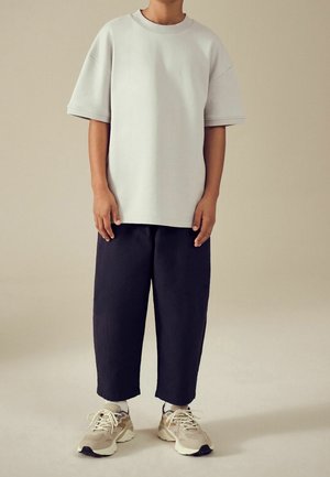 Enfant portant un T-shirt gris clair trop grand, un pantalon noir ample et des baskets beiges, debout devant un fond neutre uni.