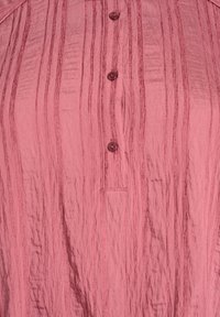 Detailný záber na ružovú textúrovanú látku s vertikálnymi pruhmi a tromi malými okrúhlymi gombíkmi usporiadanými vertikálne.