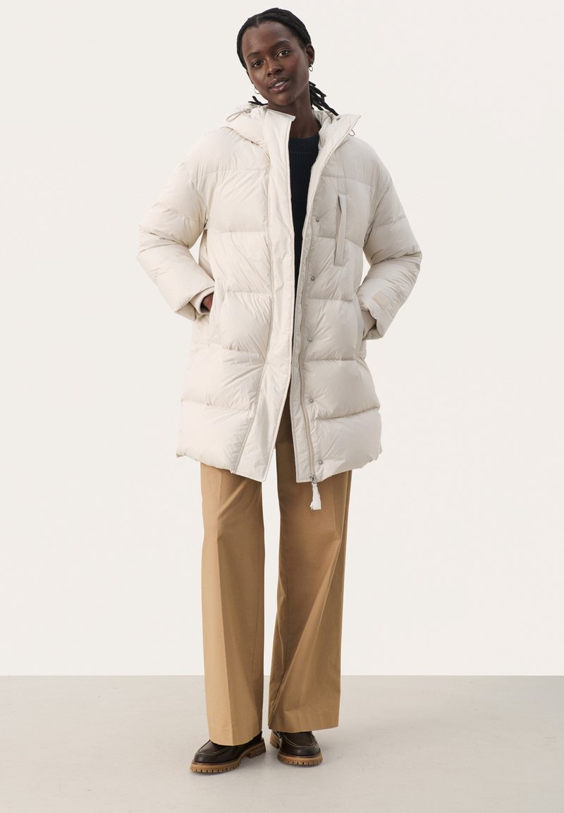 Lekka, kremowa puchówka z kapturem, z zapięciem na zamek błyskawiczny, kieszeniami bocznymi oraz pikowanym wzorem. W połączeniu z beżowymi spodniami.