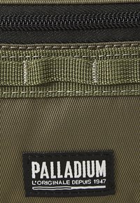 Geanta branduită Palladium, realizată dintr-un material texturat de culoare verde măslin, cu un fermoar negru și o etichetă rectangulară care afișează numele brandului.