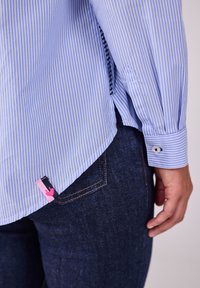 Hellblau gestreiftes Hemd aus gewebtem Stoff, mit knöpfbaren Manschetten und einem pinken Akzent-Tag am Saum. Getragen über dunklen Jeans.