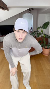 Šviesiai pilkas užtrauktukas megztinis su tamsiai pilkomis rankovėmis, kreminiai pločio kojos kelnės, balti sportbačiai ir šviesiai mėlynas Nike kepurė, pozavo viduje.