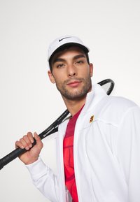 Chaqueta blanca con cremallera sobre una camiseta gráfica roja, con una raqueta de tenis negra sostenida sobre el hombro. Gorra blanca con logo en la cabeza.
