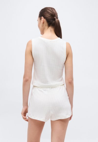 Γυναίκα με καστανά μαλλιά σε ένα χαμηλό αλογοουρά, που φοράει λευκό αμάνικο τοπ και λευκά σορτς, στέκεται με την πλάτη προς τον θεατή σε απλό φόντο.