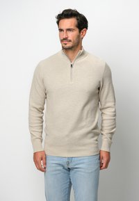 Beige zippered tröja med en strukturerad stickad mönster, långa ärmar och en figurnära silhuett, kombinerad med ljusblå jeans.