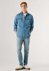 Jeune homme debout, les mains dans les poches, portant une chemise en jean bleu clair, un jean délavé, des baskets blanches et un t-shirt blanc, sur un fond blanc.