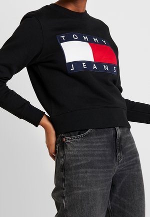 Persoon draagt een zwarte trui met een groot rechthoekig Tommy Jeans-logo en vervaagde zwarte spijkerbroek.