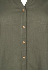 Chemise en lin vert olive avec un col en V et six boutons en bois sur le devant. Présente un tissu texturé et une couture simple.