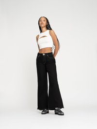 Weißes, kurzes Tanktop mit Cut-Out-Details, kombiniert mit hochgeschnittenen, schwarzen Schlagjeans. Verfügt über kontrastierende Nähte und ausgefranste Säume. Schwarze Schuhe.