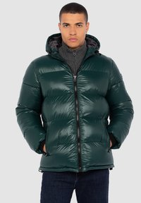 Chaqueta de plumas verde oscuro con acabado brillante, con cierre de cremallera frontal, capucha y acolchado horizontal; combinada con un suéter gris y jeans oscuros.