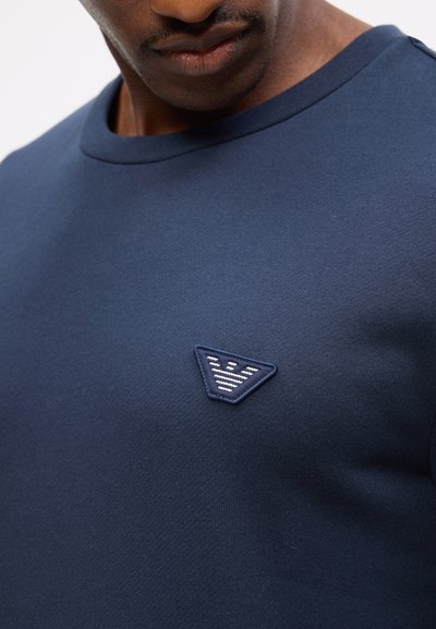 Άνδρας που φοράει μπλε σκούρο πουκάμισο με στρογγυλή λαιμόκοψη και μικρό τριγωνικό κεντημένο λογότυπο στο στήθος.