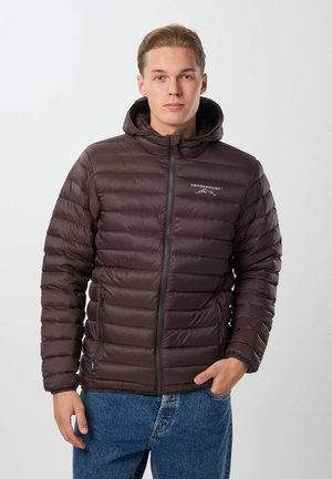 Jeune homme portant une veste matelassée marron foncé avec capuche, fermée par une fermeture éclair, et un jean bleu, debout devant un fond clair uni.