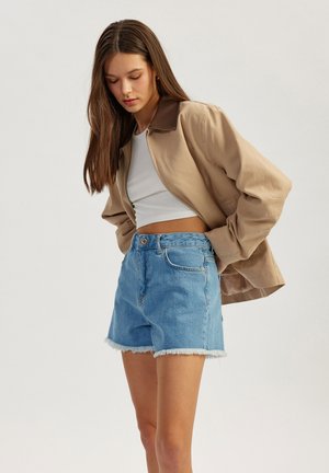 Beige bomber jacket with brown collar, white crop top, and light blue denim shorts with frayed hems. Casual, layered outfit for warm weather.