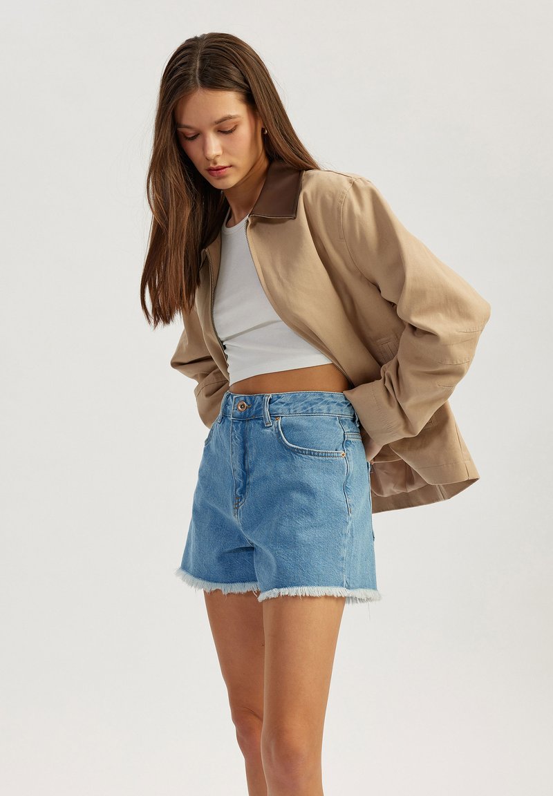 Chaqueta bomber beige con cuello marrón, top corto blanco y shorts de mezclilla azul claro con dobladillos deshilachados. Atuendo casual en capas para el clima cálido.
