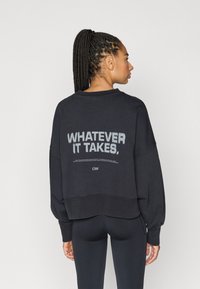 Sort sweatshirt med løs pasform, brede ærmer og kortet kant. På ryggen står der med hvid tekst "WHATEVER IT TAKES."