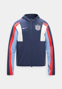 Veste de sport bleu marine à fermeture éclair avec capuche, panneaux blancs près des épaules, rayures rouges et bleu clair sur les manches, écusson de football d'Angleterre et logo Nike.