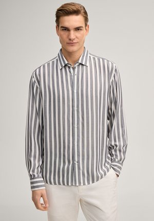 Junger Mann mit zurückgekämmtem Haar, der ein graues und weißes, vertikal gestreiftes Hemd und weiße Hosen trägt, stehend vor einem schlichten Hintergrund.