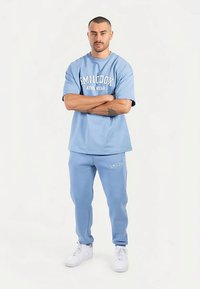 Ljusblå bomullsathleisure-outfit med en kortärmad tröja med texten "SMILODOX ATHL WEAR" och matchande joggers. Vita sneakers.