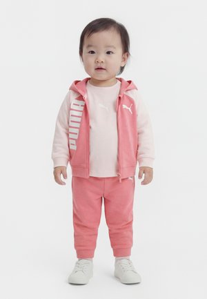 Νήπιο που φοράει ροζ φόρμα Puma με γιλέκο με κουκούλα, μπλούζα μακρυμάνικη, παντελόνι και άσπρα αθλητικά παπούτσια, στέκεται σε λευκό φόντο.