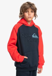 Sweatshirt à capuche bleu marine et rouge avec des manches longues et une poche frontale. Présente un logo Quiksilver texturé sur la poitrine et des poignets et un ourlet côtelés.