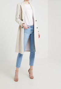 Abrigo largo beige con un diseño a medida, botones de doble botonadura, combinado con unos jeans ajustados azul claro y sandalias de tacón alto beige.