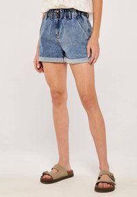 Denim-Shorts mit elastischem Bund, umgeschlagenen Säumen und sanften Ausbleichungen. Kombiniert mit flachen, offenen Sandalen in Beige.