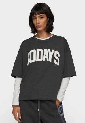 T-shirt gris surdimensionné avec "100JOURS" imprimé en blanc, doté d'un col rond et de manches courtes, superposé sur une chemise à manches longues.