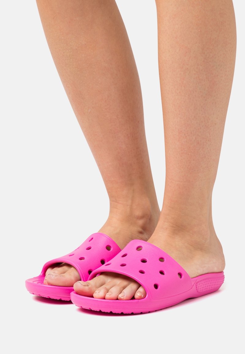 Sandales en caoutchouc roses avec des trous de ventilation, design ouvert à l'avant, et semelle texturée, montrées sur des pieds nus en position debout.