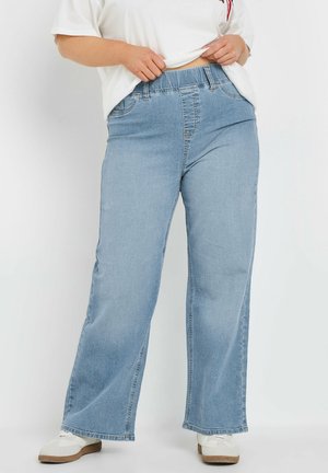 Osoba w jasnoniebieskich jeansach o szerokich nogawkach, białych trampkach i białej koszulce podnosząca koszulkę, aby pokazać pas spodnie.