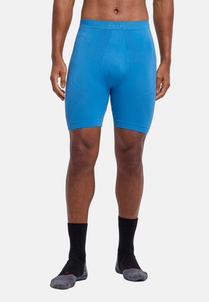 Mężczyzna w niebieskich sportowych krótkich spodenkach kompresyjnych i czarnych skarpetach, stojący na neutralnym tle.