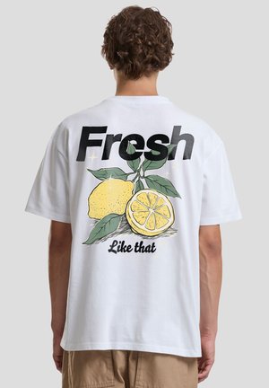 Osoba s kučeravými vlasmi otočená chrbtom, nosiaca biele tričko s grafickým motívom citróna a nápisom „Fresh“ a „Like that“ na chrbte.