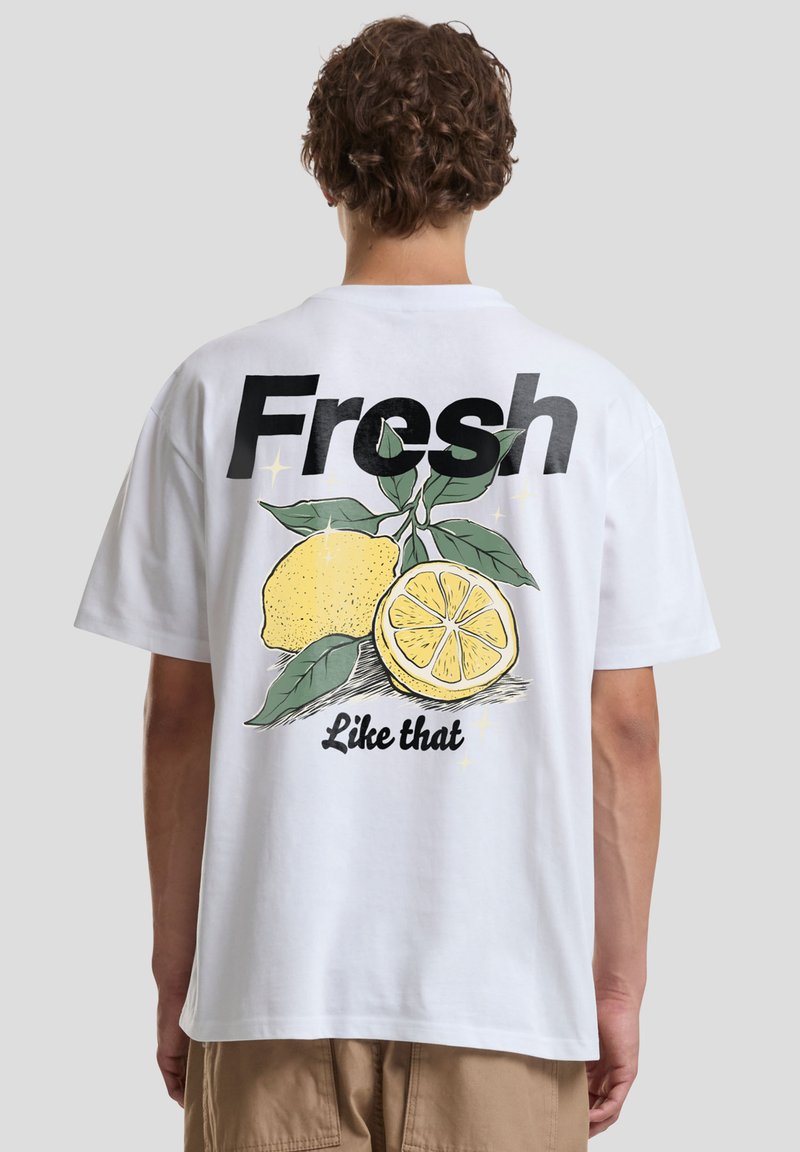 Personne aux cheveux bouclés, vue de dos, portant un T-shirt blanc avec un motif citron et les textes « Fresh » et « Like that » au dos.