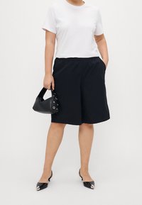 Witte T-shirt met korte mouwen gecombineerd met zwarte shorts met elastische taille. Zwarte handtas met ronde uitsparingen en glanzende zwarte instappers.