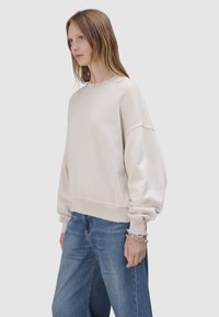 Młoda kobieta o jasnobrązowych włosach ma na sobie oversize'ową kremową bluzę, luźne niebieskie dżinsy i wiele srebrnych bransoletek na lewej ręce na szarym tle.