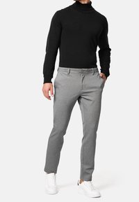 Homme portant un col roulé noir, un pantalon gris slim et des baskets blanches, debout avec une main dans la poche devant un fond blanc.