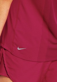 Vinröd nät sporttröja och shorts som bärs av en person, med en liten reflekterande Nike-logga nära tröjans fåll.
