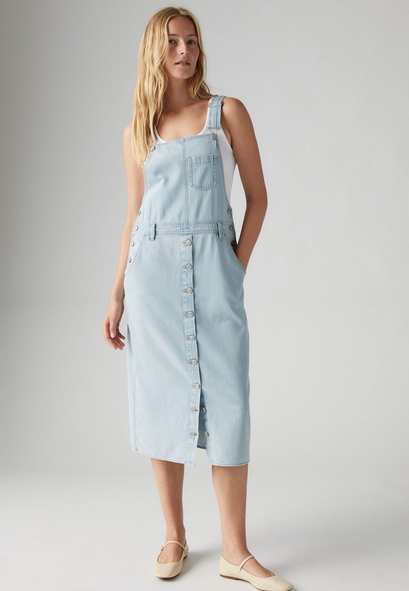 Denim-Latzkleid in Hellblau, mit verstellbaren Trägern, Fronttasche, durchgehender Knopfleiste und Seitentaschen. Glatte Textur und Midi-Länge.