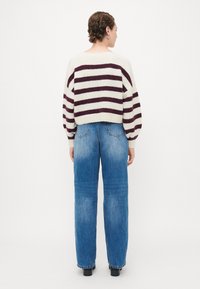Person steht mit dem Rücken und trägt einen weiß-rot gestreiften Pullover, hochtaillierte blaue Jeans und schwarze Schuhe.
