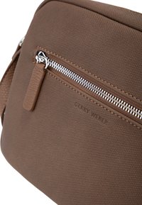 Hnědá syntetická crossbody taška s texturovaným povrchem, stříbrným zipem a koženým poutkem, s vyraženým názvem značky.