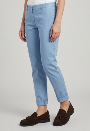 Světle modré kalhoty s manžetami, oblečené osobou stojící, doplněné tmavě hnědými mokasíny s třásněmi na jednobarevném pozadí.