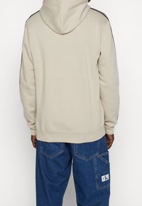 Person i beige hoodie med svarta ränder på ärmarna och blå denimjeans med en sidoficka märkt CK, visas från baksidan.