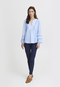 Világoskék, hosszú ujjú, V-nyakú felső, gyűrött részletekkel és hímezett díszítéssel; sötétkék, slim-fit nadrággal és bézs mokaszinokkal párosítva.