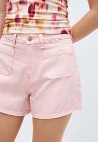 Ροζ ανοιχτόχρωμο σορτς τζιν με εμπρόσθιες τσέπες και κλείσιμο με κουμπί, φορεμένο με αμάνικη πολύχρωμη μπλούζα με ρυτιδώσεις.